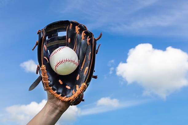 A black baseball glove with a white baseball inside, held against a blue sky.